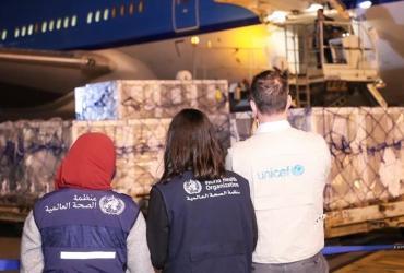 Three people wearing vests of World Health Organization and UNICEF observing shipping pallets being off-loaded from an airplane at night.