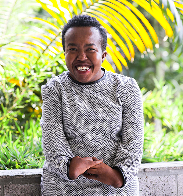 Liliane Akdata, smiling warmly, sits outside wearing a long-sleeved shirt with a black and white horizontal zig-zag pattern. It is such a sunny day that the greenery behind her seems to glow.