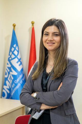 A smiling woman standing with her arms crossed. Flags of Iraq and UN Women show in the background.