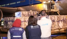 Three people wearing vests of World Health Organization and UNICEF observing shipping pallets being off-loaded from an airplane at night.