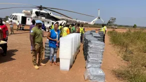 UN Volunteer Angeline Ambogo Kidiga, wearing a blue vest, stands beside stacks of electoral materials being unloaded from a UN helicopter, coordinating logistics and operations in Kaga Bandoro.