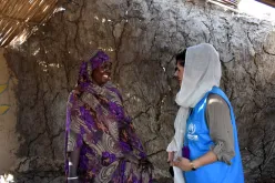 A woman wearing a blue vest (on the right) standing face-to-face to a woman wearing a traditional dress. Both women are smiling at each other. A clay wall appears in the background with a hay roof above.