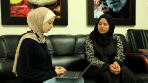 Two women seated indoors on a couch. The young woman to the left is a UN Volunteer typing on her laptop while interviewing the other woman, who is a beneficiary from one of UN Women programmes in Gaza, State of Palestine.