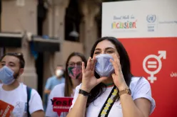 A young woman wearing a face mask shouting with her hands held to the sides of her mouth. In the background, two young people with face masks holding signs, and a banner showing the word inclusion and the logo for the Sustainable Development Goal 5 on gender equality.