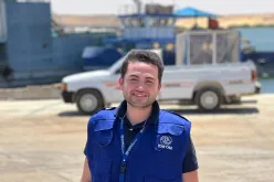 A young man wearing a blue vest with the logo of the International Organization for Migration standing at a border crossing point. A pick-up vehicle and a docked ship appear in the blurry background.