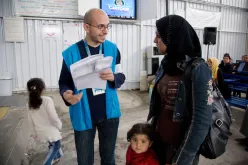 A man in blue vest assists a woman and her child at a refugee service center.