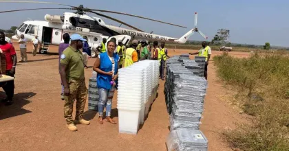 UN Volunteer Angeline Ambogo Kidiga, wearing a blue vest, stands beside stacks of electoral materials being unloaded from a UN helicopter, coordinating logistics and operations in Kaga Bandoro.