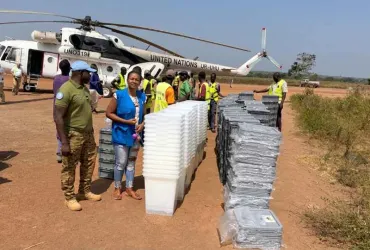 UN Volunteer Angeline Ambogo Kidiga, wearing a blue vest, stands beside stacks of electoral materials being unloaded from a UN helicopter, coordinating logistics and operations in Kaga Bandoro.