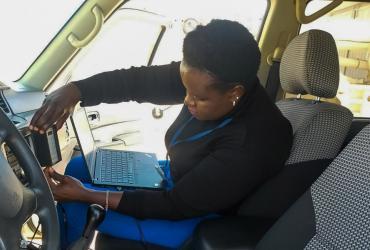 A woman sitting in a parked vehicle with an open door. She has a laptop on her lap while attaching a cable to a device fixed inside the vehicle.