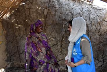 A woman wearing a blue vest (on the right) standing face-to-face to a woman wearing a traditional dress. Both women are smiling at each other. A clay wall appears in the background with a hay roof above.