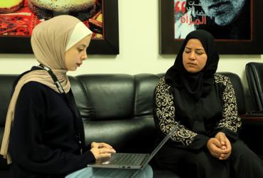 Two women seated indoors on a couch. The young woman to the left is a UN Volunteer typing on her laptop while interviewing the other woman, who is a beneficiary from one of UN Women programmes in Gaza, State of Palestine.