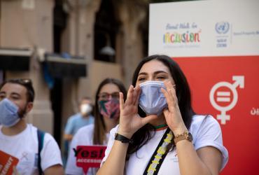 A young woman wearing a face mask shouting with her hands held to the sides of her mouth. In the background, two young people with face masks holding signs, and a banner showing the word inclusion and the logo for the Sustainable Development Goal 5 on gender equality.