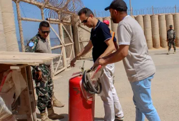 Three men standing outdoors in a walled area, inspecting a red cylindric fire extinguisher.