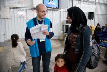 A man in blue vest assists a woman and her child at a refugee service center.