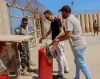 Three men standing outdoors in a walled area, inspecting a red cylindric fire extinguisher.